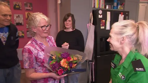 Bob Dewane left, Jackie centre and Amy in the background with Sarah Taylor on the right wearing her St John's Ambulance uniform. Jackie is handing Taylor some flowers and they are all stood in the family's kitchen.