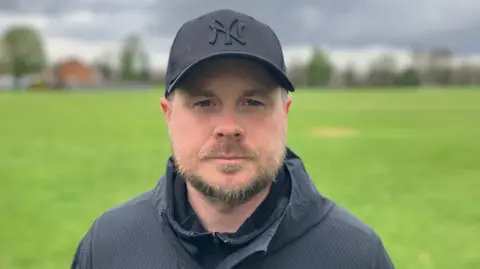 Sam Read/BBC Jamie Conroy stands in a park with a grass field. He wears a New York Yankees baseball cap with a black coat on above a black jumper.
