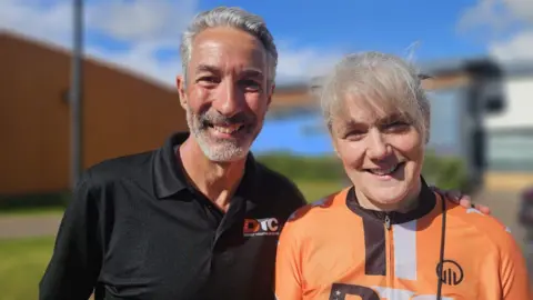 Chris Redding is smiling at the camera, he has short grey hair and is wearing a black polo top. He has his arm around Susannah Simpson who has grey hair tied back with a fringe. She is wearing an orange cycling jersey. 