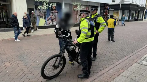 George King/BBC PC Matthew Dilks and PC Lewis Finch seizing an illegally modified e-bike in Ipswich town centre. The rider has been made anonymous and there are shoppers in the background.