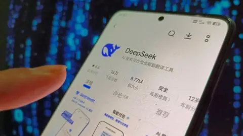 A finger hovers over a phone with the DeepSeek logo, a blue whale, on the screen.