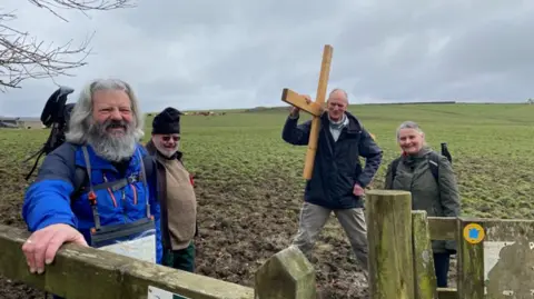 A cheerful looking man with a grey beard and hair is standing by a gate. Three other people are visible behind, one has a cross on his shoulder. It is very muddy