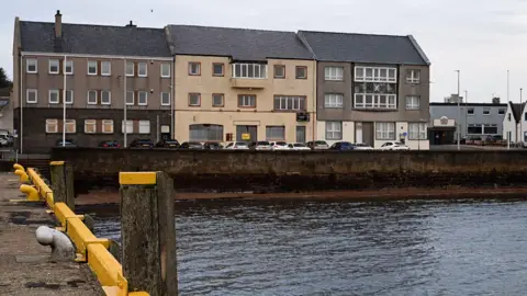 A view over part of Stornoway harbour to the Caledonian Hotel. It is three storeys tall and cars are parked at the front of the building. 