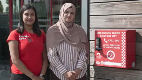 Sienna Ahir, Member of Youth Parliament and founder of Daggers Down, and councillor Obaida Ahmed stand by one of the kits at WV Active Bilston-Bert Williams. Sienna has a red t-shirt on with Daggers Down on it and Obaida has a headscarf on over a striped shirt. 