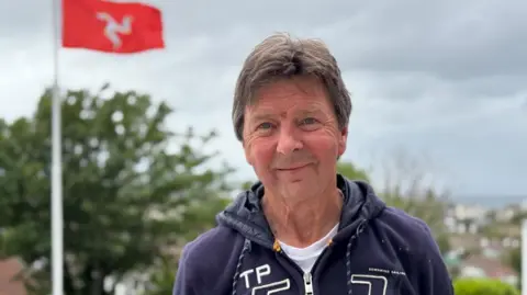 Glen Kinrade has short brown hair and is wearing a dark blue hoodie. He's standing outside with trees behind him.