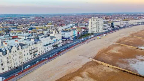 Aerial view of Eastbourne seafront. Sandy beach in the foreground and cream buildings. The sky is blue, purple and cloudy.