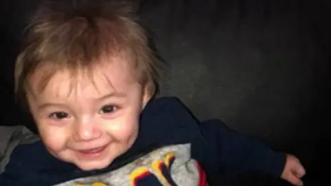 Ethan, a smiling young boy with dark hair, wearing a blue, grey and red jumper that says "Play" on it. 