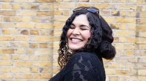 Gemma has long curled black hair and is wearing a black lace dress. She has her eyes closed and is smiling. A light yellow brick wall is in the background of the image. 