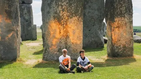 PA The Stonehenge monument on a bright sunny day. Some of the stones appear to have orange paint on them. Two of the protestors, Ms Lynch and Mr Naidu are sat in protest in front of the stones.