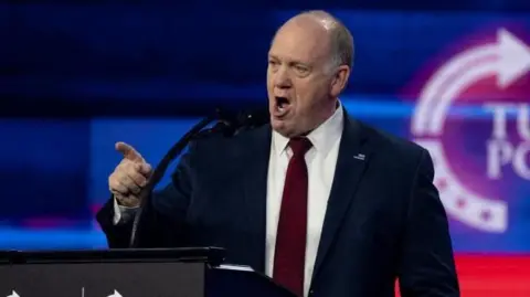 Tom Homan, wearing a suit, talks into a microphone.