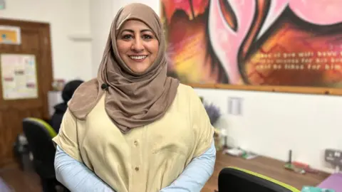 Selima is stood in the EYST office, with a large colourful piece of art on the wall behind her. She is smiling at the camera and is wearing a cream blouse and pale brown hijab.