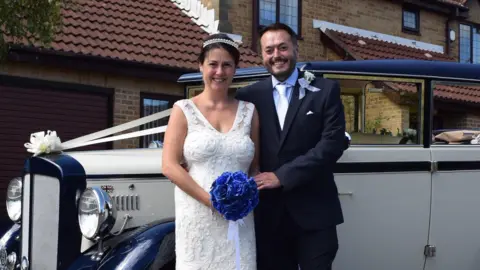 Anna-Louise Stubbings Anna-Louise and Zach on their wedding day. They are standing in front of a vintage car, Anna is wearing a white sleeveless wedding dress and holding a blue bouquet. Zach is wearing a navy suit. Both are smiling,