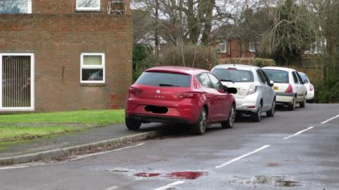 Four cars parked on a road. One car is red, the others are silver. They are parked partly on the pavement. To the left, there is a building and some grass.