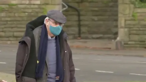 A man with short grey hair, wearing a face mask and flat cap as well as a blue sweatshirt and scarf and grey jacket. He is walking along a pavement with a road behind him.