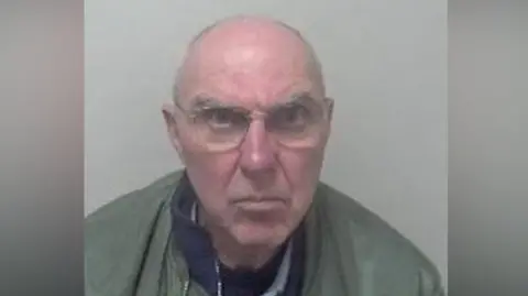 Kent Police An elderly bald man in a mugshot. He is wearing a green jacket and glasses.