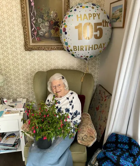 June Spencer in an armchair with a 105th birthday balloon and with a pot plant on her lap