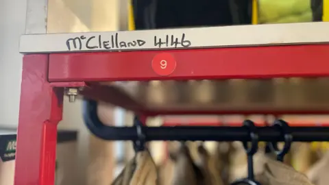 A red coat railing is pictured. It has a metal frame with the tops of some coats hanging under it to be seen. It has the number nine in the middle. Above on a white frame, the name "McClelland" and number 4146 is written in black permanent marker ink.