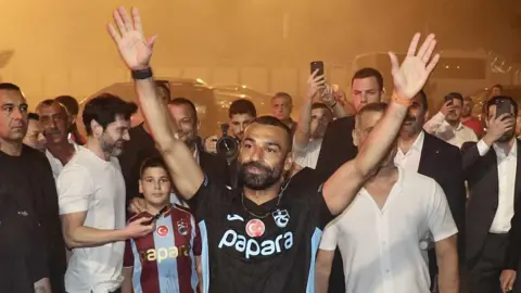 Mohamed Salah with a black Trabzonspor top on with his hands in the air outside Trabzon airport
