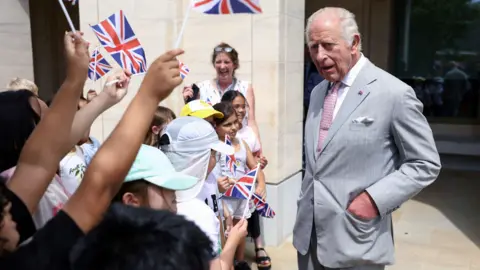 PA Media King Charles on the right wearing a grey suit and he is talking to children waving British flags.