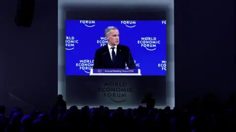Reuters Canada's Prime Minister Mark Carney appears on a screen as he speaks during the 56th annual World Economic Forum (WEF) meeting in Davos, Switzerland. The foreground of the image shows the heads of spectators illuminated by the glow from the screen, which features Carney speaking at a podium with a royal blue backdrop behind him that shows the World Economic Forum logo in white