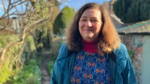 A woman, Di, smiling at the camera. She is standing in a garden, with a path visible in the background. The sun is shining.