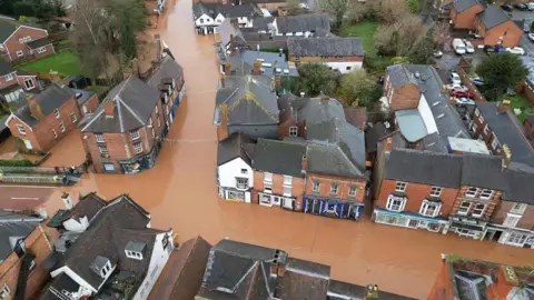 Flooded streets in Tenbury Wells. Long stretches of roads are under water, with water levels appearing several centimetres deep. The water is brown. There are no people or vehicles visible on the water. Shops, homes and businesses can be seen.