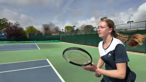 Daniella Britton in a white and navy blue t-shirt on a blue and green court holding a tennis racket and looking off camera.