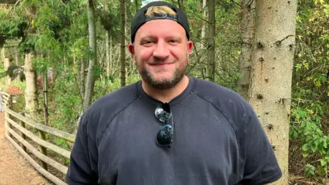 A man wearing a dark T-shirt and cap standing beside a wooden fence in a wooded area.