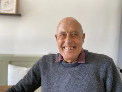 An older, bald man in a blue jumper beaming with a broad smile.