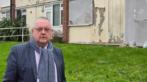 LDRS Image of Kevin Clarke who is wearing a suit jacket and a grey scarf. He is stood in front of some dilapidated houses. 