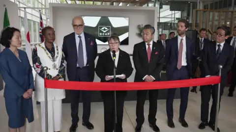 A row of dignitaries standing in a row ahead of the opening of the ceremony on Monday, with them standing in a line next to three flags - an Italian flag, a Japanese flag and the Union flag. A picture of the jet is behind the people.