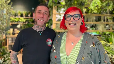 A man with short brown hair is wearing a black t-shirt and has tattoos on his neck and arm. He is standing next to a woman with red hair and black framed glasses, she is wearing a green gingham top and a cardigan with bug prints. They are standing in their plant shop with plants surrounding them.