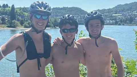 Three 18 year olds are stood shoulder to shoulder with cycling helmets, glasses and their tops off on a hot sunny day, in front of a large lake and countryside behind them. They are looking at the camera and smiling.