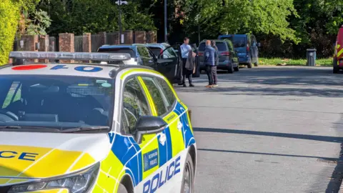PA A marked Metropolitan Police car is parked on a residential street in north‑west London while emergency vehicles and several people stand beside parked cars further up the road, with trees and brick walls lining the street.