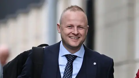 Jamie Bryson walking outside of court, he is smiling and wearing a navy three-piece suit with a light blue shirt and a navy tie with white dots. He has short light hair and stubble and is wearing a black backpack. 