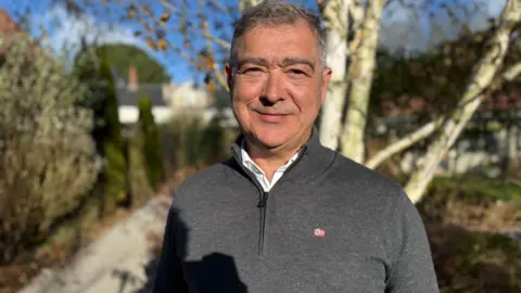 BBC A man is standing outdoors, looking at the camera and smiling. He is wearing a grey jumper. There are trees and bushes with a path in the background. The sky is blue. 