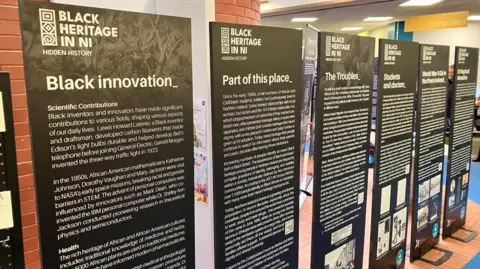 Six black signposts with white writing are stood side-by-side. 
They read from left to right: 'Black innovation, Part of this place, The Troubles, Students and Doctors, World War II GIs in Northern Ireland'. 