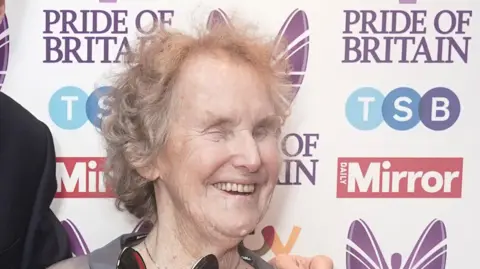 Jill Allen-King has short grey hair and is smiling at an awards evening. A board covered in the event's sponsors is behind her.