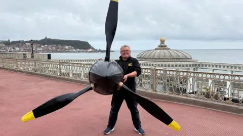 A man dressed in black on the seafront with the propellor from a replica Mk1 Spitfire in his hands