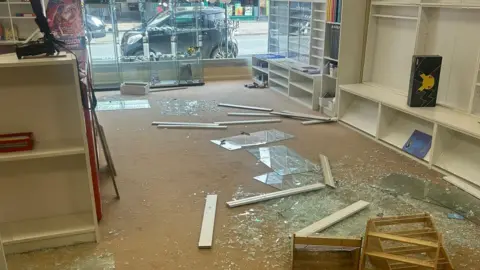 Smashed glass and broken shelving covers the floor of Trove indoor market in Winton. The shop window looks out on to a street with a parked car outside.