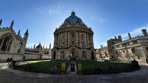 The Radcliffe Camera on a sunny day. The building itself is in shadow and is backlit against a blue sky. Other historical buildings can be seen around it, with a round lawn and cobbled street between them. 