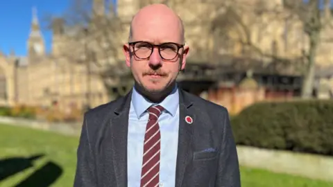 A bald man with glasses outside the Westminster Parliament on a sunny day. He is looking at the camera and is wearing a grey suit, blue shirt and maroon and grey striped tie. 