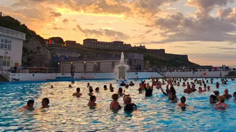 The lido is full of swimmers as the sun nearly rises above the surrounding buildings. Three of the swimmers have their arms raised above their heads. There is a fountain in the lido and the clouds above are pink and orange.