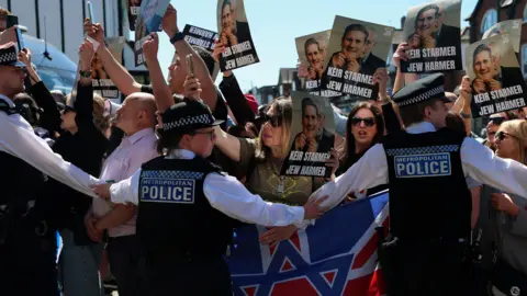 Three police officers stand in front of a group of protesters holding signs reading: "Keir Starmer Jew Harmer". The placards also depict a picture of former Labour leader Jeremy Corbyn holding a mask of Keir Starmer in front of his face. 