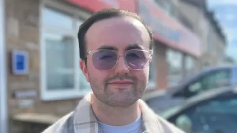 A close-up of Jordan Robbins, who has short hair and a short beard. He is wearing tinted glasses and a white t-shirt with a checked shirt over the top. The blurred background features a building and two parked cars.