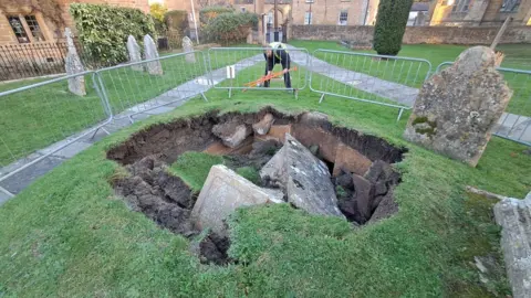 A stone box tomb which has collapsed into a large hole in the ground, through the soil and into the crypt below, a small stone home buried underground. There are metal barriers around the hole and a man in a high-vis jacket. There are other gravestones very close to the edge of the hole, which is about 12ft (3.6m) wide.