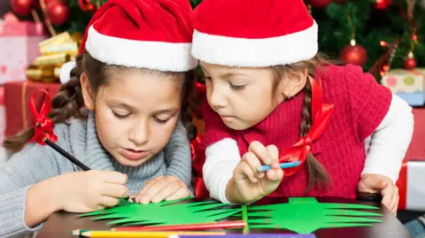 Getty Images Two young girls wearing red and white Santa hats are colouring green paper Christmas trees in front of a large Christmas tree. Both have pencils in their hands and one is looking at the other's design.