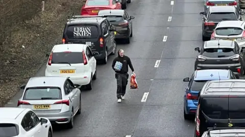 A man in a black hoodie walks down a stretch of road between queues of traffic. He has a large bag of crisps in his hand and bottled water. 