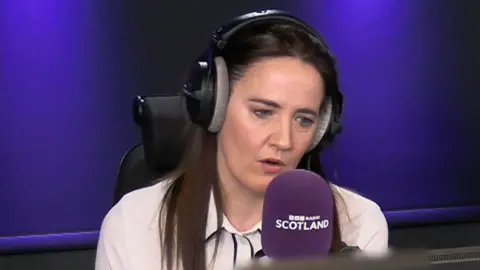 A woman with long dark brown hair is speaking into a purple microphone that reads 'BBC Radio Scotland'.
