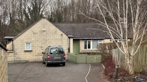 Newton House in Bath. It looks like a residential bungalow set back from the road with a large driveway. The home is made of light-coloured bricks and dark roof tiles, with a mint-green picket fence and front door.  The home is surrounded by trees which have been stripped of their leaves in winter. 